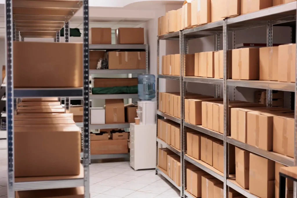 Armazenamento de caixas e papelões em prateleiras metálicas de um estoque, com corredores organizados e materiais de embalagem prontos para envio.