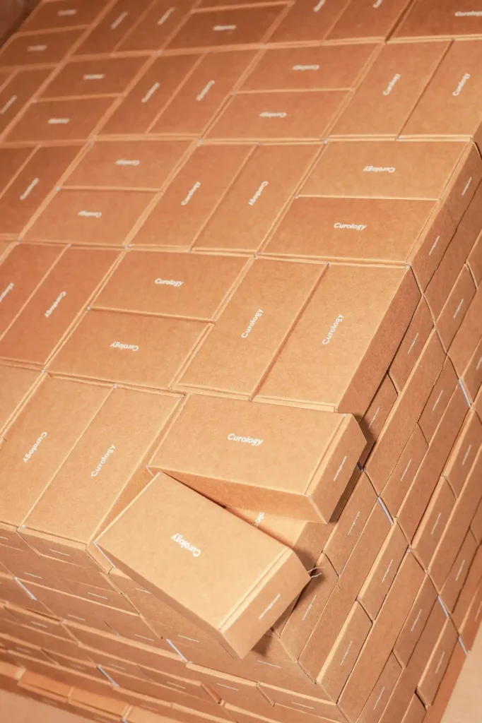 Caixas de papelão empilhadas com a marca “Curology”, em um ambiente de armazenagem, com destaque para os volumes alinhados e fitas de amarração.