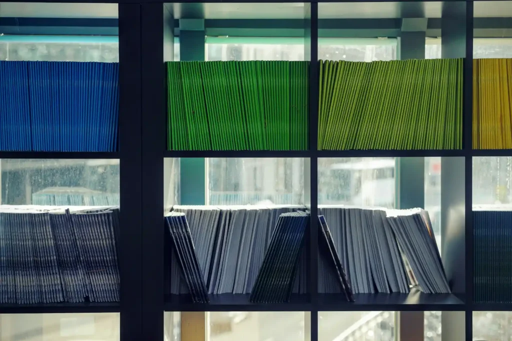 Molduras de vidro ou placas transparentes com painéis ondulados em diferentes cores (azul, verde, amarelo), organizadas em prateleiras, com vista através de uma estrutura escura.