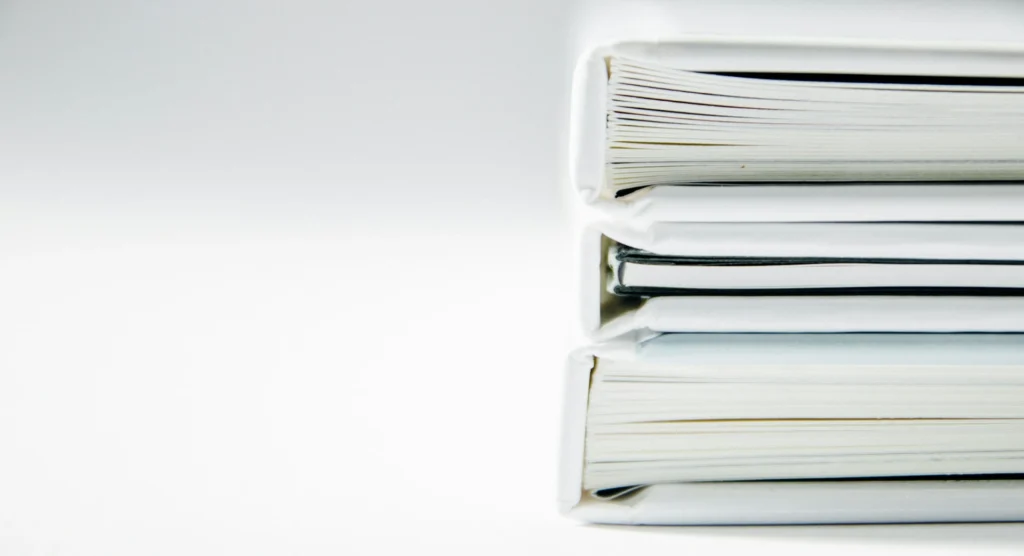 Pilhas de livros de capa branca empilhados, mostrando as lombadas e páginas fechadas em fundo claro, com foco no conjunto de volumes organizados.